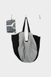 The Indigo Link tote, now available at Pierre Lafond, hangs on a wall hook, its black and gray color blocks complementing a compact patterned zip pouch beside it—both displayed against a white background.