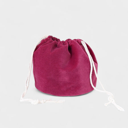 The Poppy Bag Fuchsia Velvet is a round velvet drawstring pouch with white cords, shown against a plain light gray background.