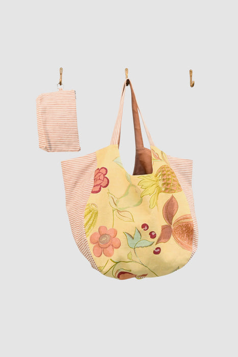 The Coral Stripe large tote bag, crafted from upcycled designer upholstery featuring pink and yellow florals and stripes, hangs on a wall hook with its matching pouch on a nearby hook.