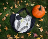 A reversible Bella Bag, available at Pierre Lafond, featuring a modern embroidered botanical print, is placed on grass amid autumn leaves beside an orange pumpkin.