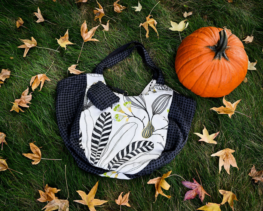 A reversible Bella Bag, available at Pierre Lafond, featuring a modern embroidered botanical print, is placed on grass amid autumn leaves beside an orange pumpkin.