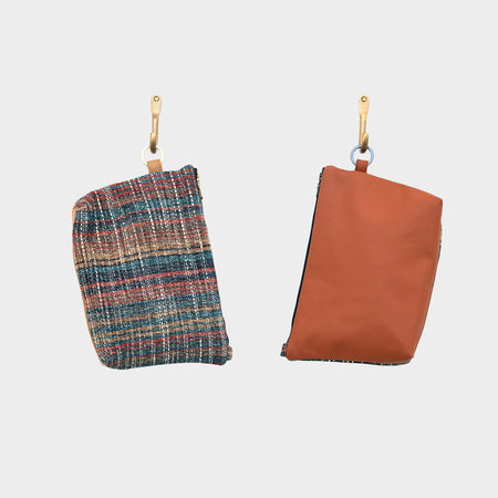 Two small pouches hang on light hooks: a colorful plaid upcycled designer pouch and Rust Horizon, a plain rust leather pouch with a visible zipper. Both feature tan straps attached to silver rings.