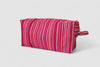 The SB Bag- Raspberry Stripe is a rectangular bag with pink, red, and purple vertical stripes on designer upholstery fabric. It has a gold zipper, small side handle, upcycled cotton lining, and is shown against a white background.