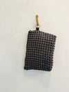 A textured, dark gray zippered pouch from AVAILABLE AT PIERRE LAFOND, crafted with upcycled designer upholstery, hangs on a gold hook against a plain white wall.