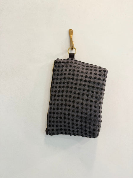 A textured, dark gray zippered pouch from AVAILABLE AT PIERRE LAFOND, crafted with upcycled designer upholstery, hangs on a gold hook against a plain white wall.