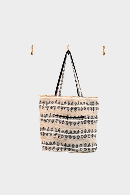 The Fern and Clay tote from AVAILABLE AT ABADIR, featuring indigo leaf block print, black handles, and a camel leather base, hangs on a wooden wall hook amid two empty hooks against a plain white background. Contact for details.