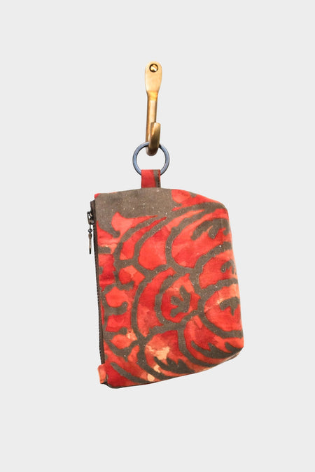 The Mini Mouse Pouch- Maori Gold is a compact essentials bag crafted from designer upholstery fabric in a red and black abstract pattern. This small square pouch has a zipper, hangs by a metal ring from a brass hook, and sits on a light background.