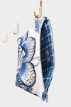 The Blue Garden pouch, now available at Pierre Lafond, hangs partially open from a hook against a plain light background, displaying its blue floral design and plaid lining. Two empty hooks are seen behind it.