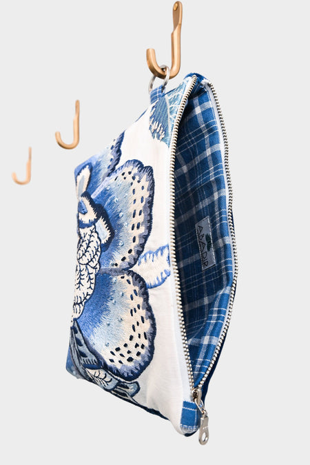 The Blue Garden pouch, now available at Pierre Lafond, hangs partially open from a hook against a plain light background, displaying its blue floral design and plaid lining. Two empty hooks are seen behind it.