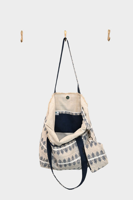 The Fern and Clay tote—AVAILABLE AT ABADIR, contact for details—features a beige and navy linen-cotton blend with a plant pattern. It hangs open on a center wall hook, flanked by two empty hooks against a plain white background.