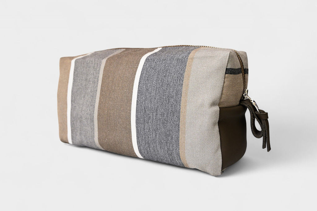 The SB Bag - Sandstone Stripe is a rectangular bag with vertical beige, gray, and brown stripes, a zipper closure, and a dark brown upcycled leather pull tab, displayed on a plain white background.