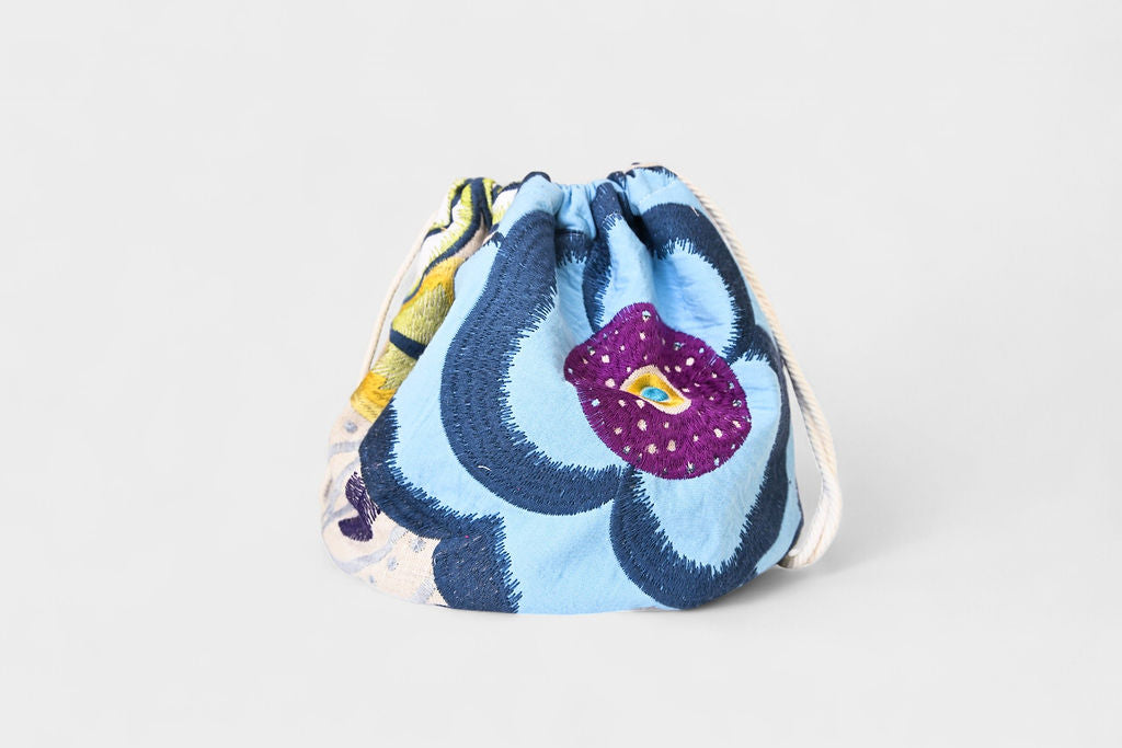 The Poppy-Fiesta Bloom Blue A is a stylish reversible drawstring bag featuring a colorful abstract floral pattern in blue, purple, and yellow. Cinched at the top, it’s perfect for everyday use and displayed on a light gray background.